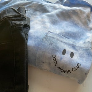 May include: A blue tie-dye t-shirt with a pocket on the front. The pocket has a black smiley face with the text "Cool Moms Club" underneath.