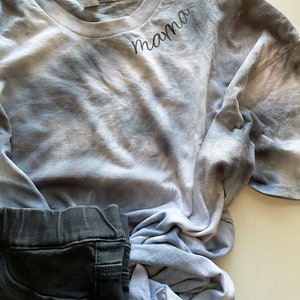 May include: A blue and white tie-dye t-shirt with the word "mama" embroidered on the front. The shirt is folded and lying on a white surface.