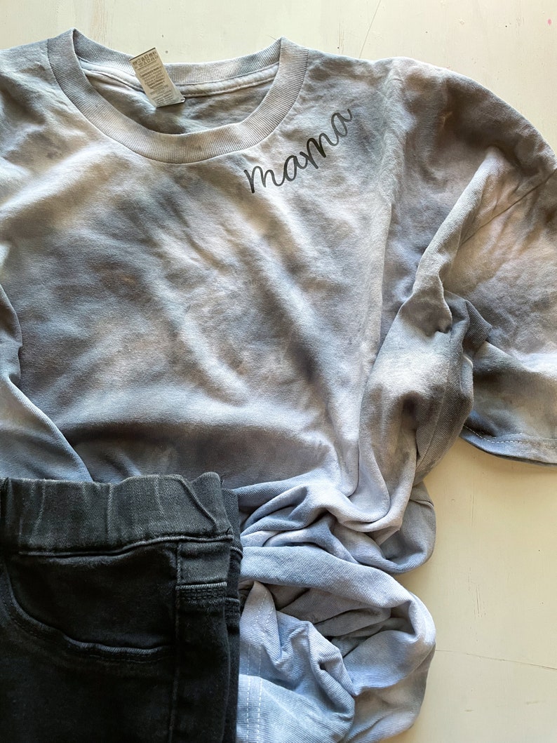May include: A blue and white tie-dye t-shirt with the word "mama" embroidered on the front. The shirt is folded and lying on a white surface.