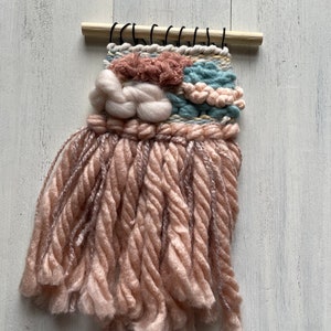 May include: A small, handmade woven wall hanging. The piece features a wooden dowel at the top with black string loops. The woven design incorporates various textures and colors, including pink, blue, and cream-colored yarn, with a long fringe.