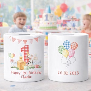 May include: Two white ceramic money banks. One features a red gingham number 1, farm animals, and the text "Happy 1st Birthday Charlie." The other has a cluster of plaid balloons and the date "26.02.2025" in red. Party decorations are in the background.