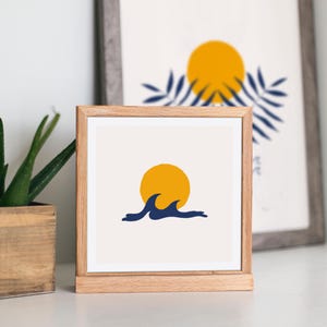 May include: A framed print with a minimalist design featuring a yellow sun setting over blue waves. The print is in a light wood frame.