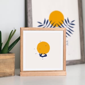 May include: A framed print of a minimalist illustration of a person diving into a large yellow sun with a few small blue birds flying around. The print is in a light wood frame.