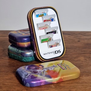May include: A vertical Nintendo 3DS game cartridge holder featuring a white interior insert with six slots for game cards. The exterior is a metallic Pokémon-themed tin with a Mewtwo graphic. Two additional Pokémon tins are stacked nearby.