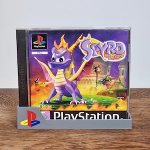 Peut inclure: Un boîtier de jeu vidéo Spyro the Dragon pour la console PlayStation, illustré par un dragon violet sur la couverture, présenté dans un support en plastique gris avec un logo PlayStation coloré sur le coin avant gauche de la base.