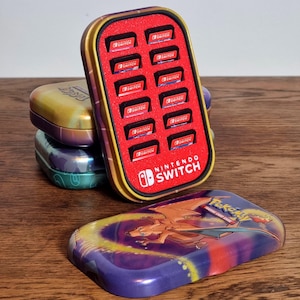 May include: A rectangular metal storage case for Nintendo Switch game cartridges with a red foam interior holding twelve game cards. The case features the Nintendo Switch logo in white. A separate metal lid displays a colourful Charizard Pokemon graphic.