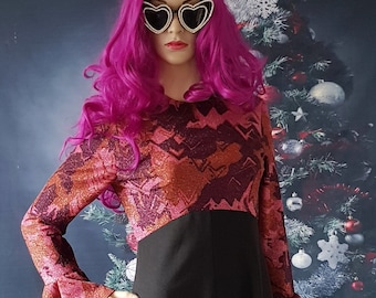 May include: A mannequin wearing a long black dress with a patterned bodice and bell sleeves. The mannequin has a pink wig, heart-shaped sunglasses, and is holding a small, beaded purse. The background features a Christmas tree and wrapped gifts.