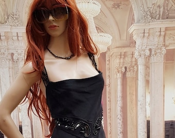 May include: A black floor-length gown with a beaded embellishment at the waist. The gown has a scoop neckline and a fitted bodice. The model is wearing sunglasses and a necklace.