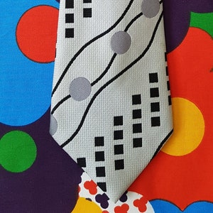 Może przedstawiać: Szary krawat z geometrycznym wzorem w kolorze czarnym i białym. Wzór zawiera czarne kwadraty, szare koła i czarne faliste linie.