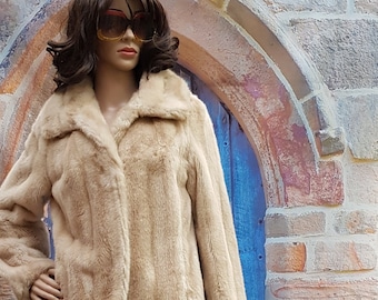 May include: A beige faux fur jacket with a brown lace skirt. The model is wearing sunglasses and standing in front of a stone archway with a blue door.
