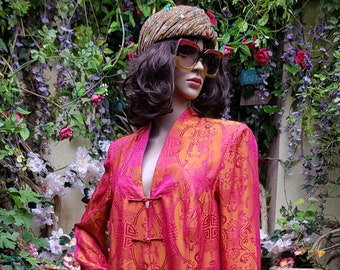 May include: A long, pink and gold robe with a traditional Chinese design and multiple gold buttons. The robe is worn by a mannequin with dark hair and sunglasses, standing in front of a wall of green plants.