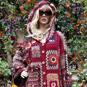 Könnte beinhalten: Ein langer, roter Häkelcardigan mit Kapuze, mit bunten Patchwork-Quadraten und Fransen. Der Cardigan wird von einer Puppe mit blonden Haaren und Sonnenbrille getragen.