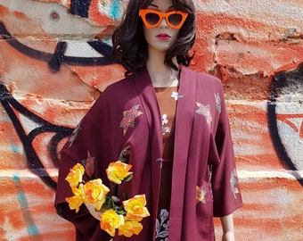 May include: A mannequin wearing a burgundy kimono with floral embroidery, over a brown dress with white floral print. The mannequin is wearing orange sunglasses and holding yellow roses. The background is a graffiti-covered wall.