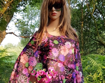 May include: A long, flowing dress with a floral pattern in shades of pink, purple, and white. The dress has long, bell sleeves and is worn by a mannequin. The background is a natural outdoor setting with trees and foliage.