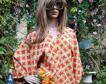 May include: A red and yellow patterned shirt with a wide collar and long sleeves. The shirt is worn by a mannequin with long brown hair and sunglasses. The mannequin is standing in a garden setting.
