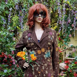 Puede incluir: Una chaqueta de ante marrón con bordados florales y una falda de estampado paisley. La chaqueta tiene una silueta larga y ajustada y un cierre de botones. La falda es de longitud maxi y tiene un esquema de color azul y verde.