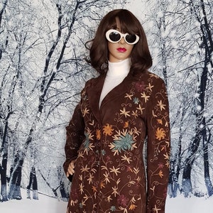 Puede incluir: Una chaqueta de ante marrón con bordados florales de colores. La chaqueta tiene una silueta larga y ajustada y un cuello. La modelo lleva la chaqueta con un jersey de cuello alto blanco y unos vaqueros azules.