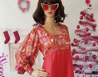 May include: A long, pink dress with a paisley print top and a solid pink bottom. The dress has long sleeves and a scoop neckline. The model is wearing heart-shaped sunglasses and a gold clutch.