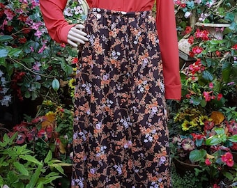 Könnte beinhalten: Eine Schaufensterpuppe trägt ein korallenfarbenes, langärmliges Hemd mit Knöpfen und einen langen Rock. Der Rock hat einen dunkelbraunen Hintergrund mit einem Blumenmuster in Orange, Weiß und Lila. Die Schaufensterpuppe trägt eine Sonnenbrille.