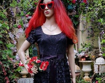 May include: A black lace dress with a ruffled skirt and a black satin sash. The dress is on a mannequin with red hair and heart-shaped sunglasses.