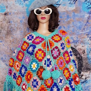 May include: A colorful, handmade crochet poncho featuring a vibrant floral pattern in shades of red, orange, blue, and purple. The poncho has a turquoise border with fringe and a drawstring with turquoise pom-poms. The garment is displayed on a mannequin.