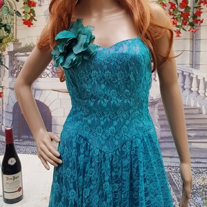 May include: A teal blue lace dress with a sweetheart neckline and a large green flower embellishment on the bodice. The dress is on a mannequin with red hair and sunglasses.