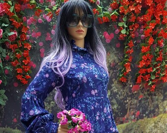 May include: A long-sleeved, blue maxi dress with a floral print. The dress has a high neck and long sleeves with ruched detailing. The dress is worn by a mannequin with purple hair and sunglasses.
