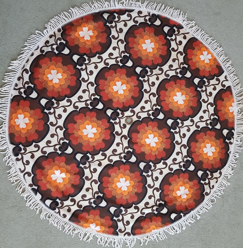 May include: A round, white, fringed tablecloth with a brown, orange, and white floral pattern.