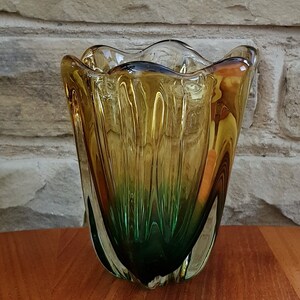 May include: A green and amber glass vase with a wavy, organic shape. The vase is made of clear glass with a green and amber color gradient.
