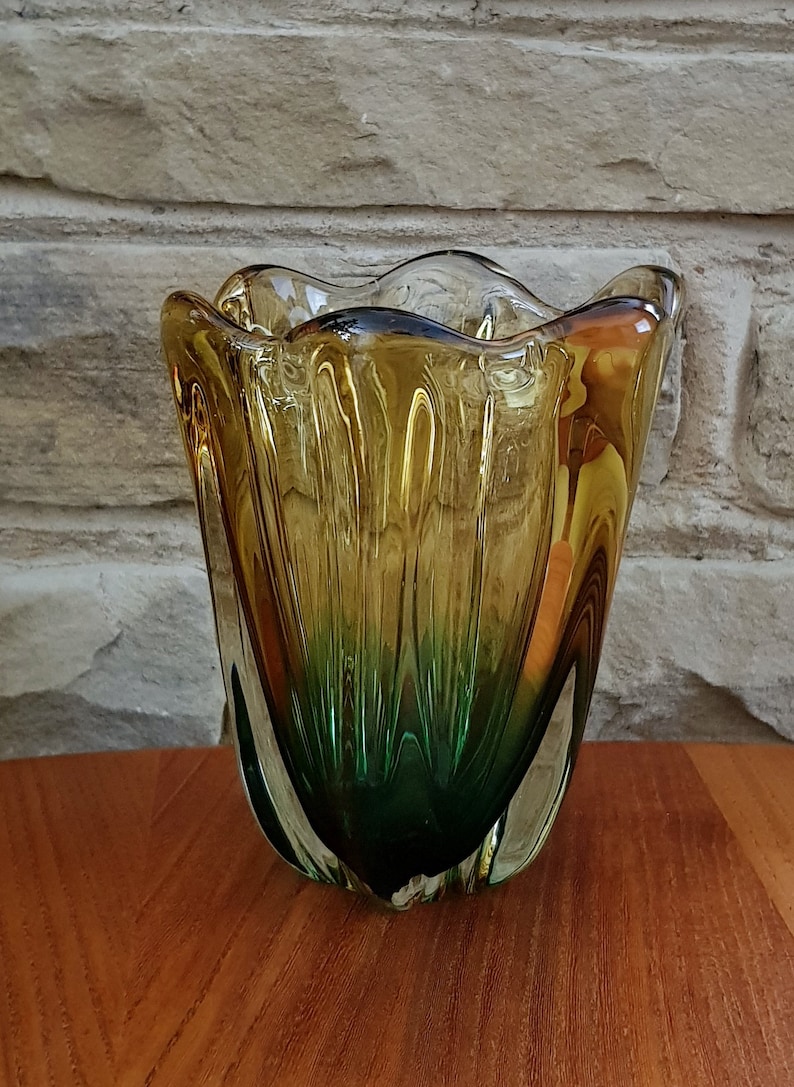May include: A green and amber glass vase with a wavy, organic shape. The vase is made of clear glass with a green and amber color gradient.