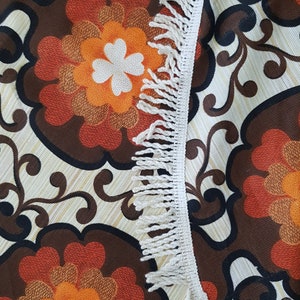 May include: A close-up of a fabric with a brown, orange, and white floral pattern. The fabric has a white fringe border.