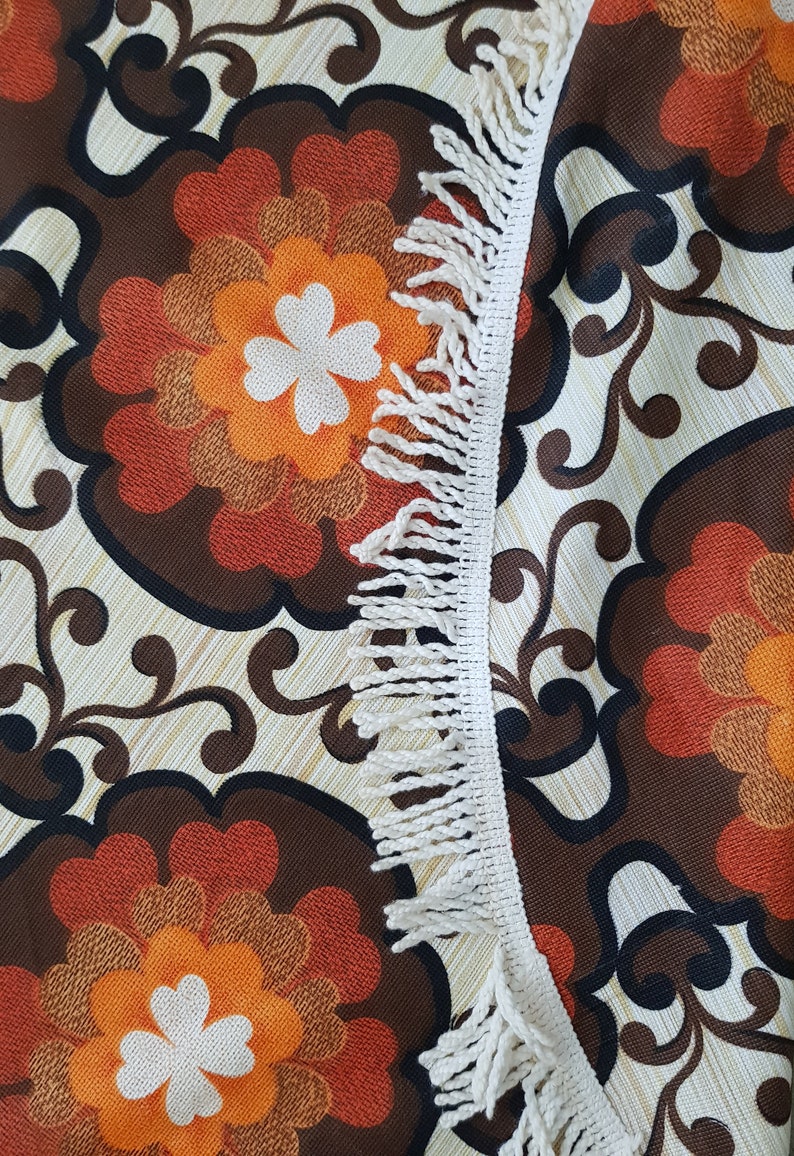 May include: A close-up of a fabric with a brown, orange, and white floral pattern. The fabric has a white fringe border.