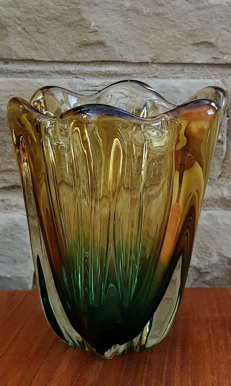 May include: A green and amber glass vase with a wavy, organic shape. The vase has a textured, rippled surface.