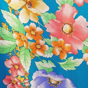Op de afbeelding: Een turquoise blauwe stof met een bloemenpatroon. De bloemen zijn in tinten geel, oranje, roze en paars. De stof heeft een gouden metallic ontwerp.