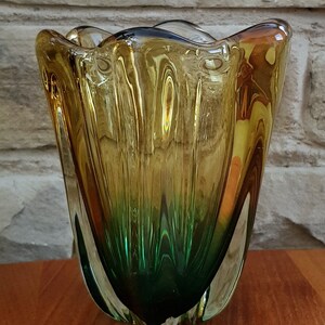 May include: A glass vase with a wavy, organic shape. The vase is made of clear glass with a gradient of amber and green colors. The vase is about 8 inches tall.
