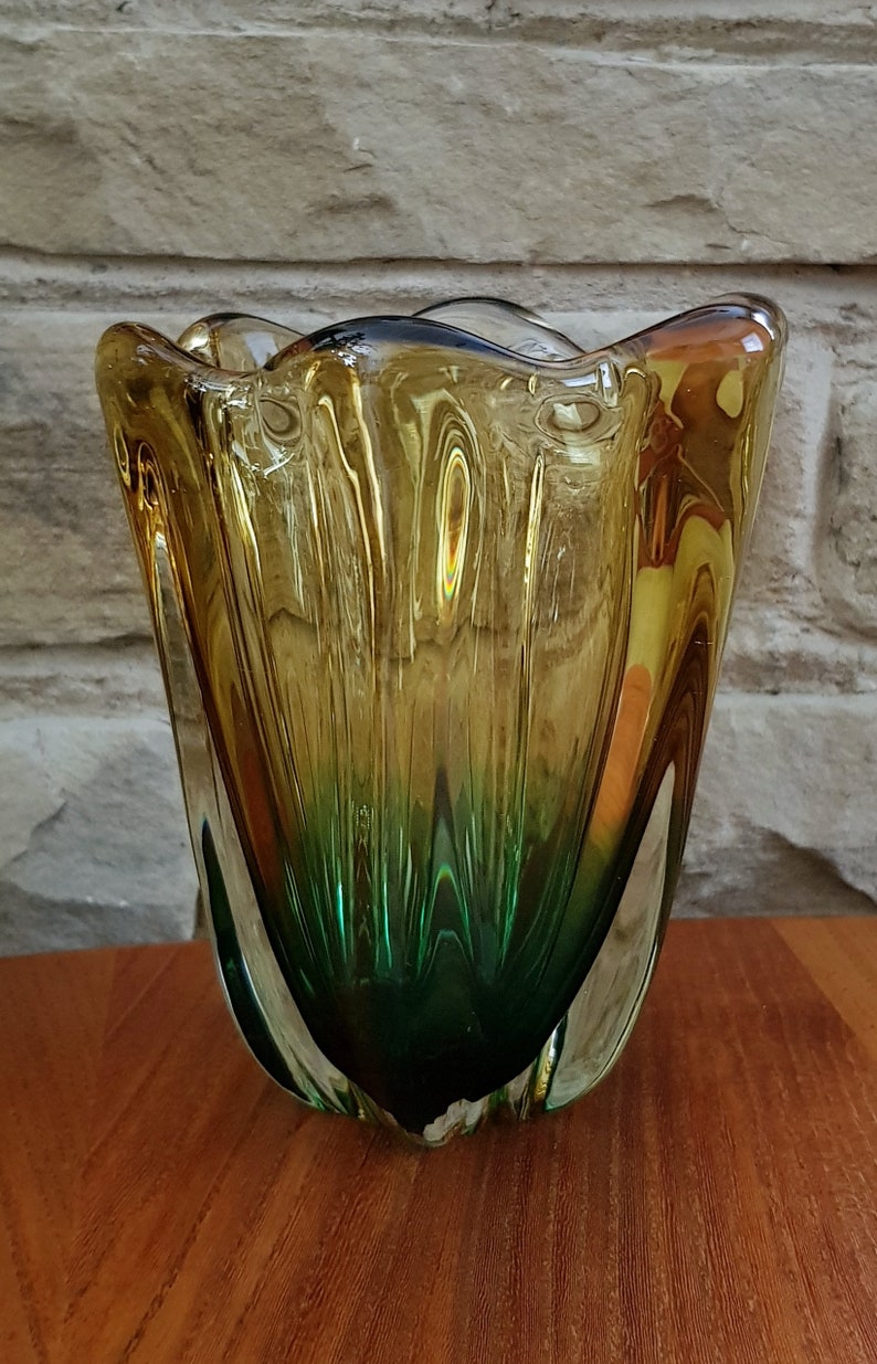 May include: A glass vase with a wavy, organic shape. The vase is made of clear glass with a gradient of amber and green colors. The vase is about 8 inches tall.