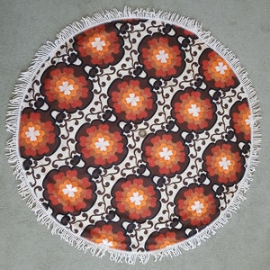 May include: A round, white, fringed tablecloth with a brown, orange, and white floral pattern. The tablecloth has a retro, 70s style.
