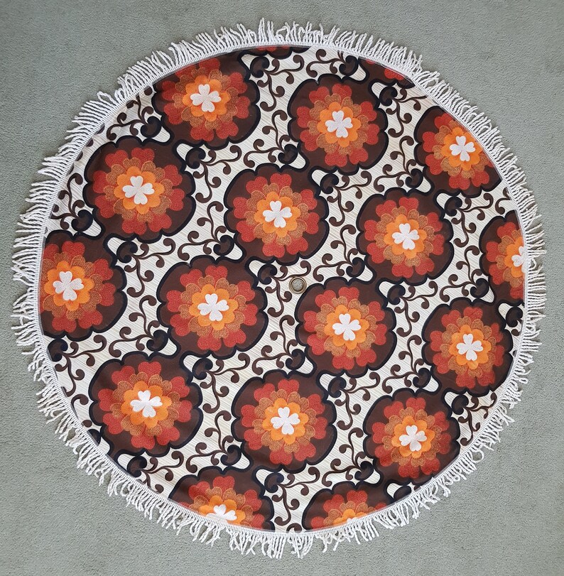 May include: A round, white, fringed tablecloth with a brown, orange, and white floral pattern. The tablecloth has a retro, 70s style.