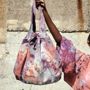 May include: A tie-dye tote bag in shades of pink, purple, and orange. The bag has a drawstring closure and long straps. The bag is held up against a stone background.