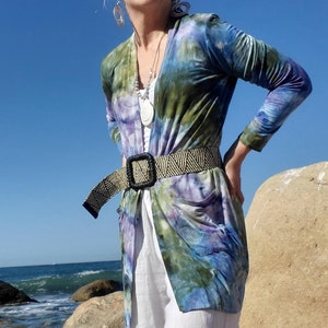 May include: A woman wearing a tie-dye cardigan with a black belt, white pants, and sunglasses standing on a rocky beach.