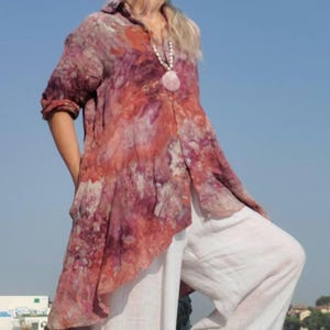 May include: A tie-dye patterned tunic in shades of red, pink, and purple, paired with white wide-leg pants. The outfit is accessorized with a long necklace and large earrings. The background features a body of water and a clear blue sky.