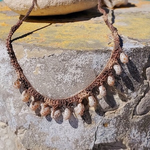 May include: A brown crocheted necklace with white seashells hanging from it. The necklace is lying on a gray concrete surface with a blurred background of a beach and blue water.