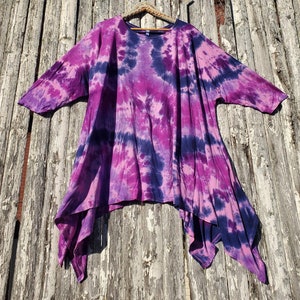 Peut inclure: Un chemisier tie-dye violet et bleu avec une longue ligne d'ourlet fluide. Le chemisier est suspendu à une clôture en bois.