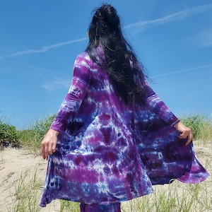 May include: A long-sleeved, knee-length duster cardigan featuring a vibrant tie-dye pattern in shades of deep purple, magenta, and blue. The garment has an open front design, a rounded hemline, and is constructed from a lightweight, flowing fabric.