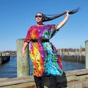 May include: A person wearing a colorful tie-dye maxi dress with a black belt and black boots. The person is standing on a wooden dock with a body of water in the background.