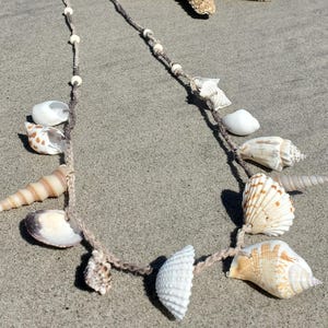 Puede incluir: Un collar de conchas hecho a mano con una variedad de conchas y pequeñas cuentas ensartadas en un cordón marrón. Una estrella de mar descansa sobre la arena junto al collar. Las conchas son en tonos blancos, beige y marrón.