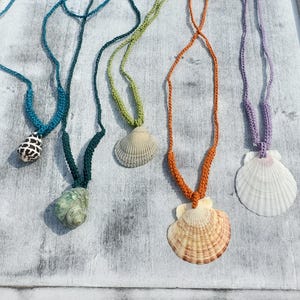 Puede incluir: Cinco collares de conchas marinas hechos a mano con cordones de ganchillo en varios colores: turquesa, verde, naranja y morado. Cada collar presenta un colgante de concha diferente, mostrando patrones y texturas naturales. Los collares se exhiben sobre una superficie blanca y desgastada.