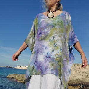May include: A tie-dye tunic in shades of blue, purple, and green, paired with a white skirt. The tunic has a V-neck and wide sleeves. The outfit is worn outdoors near the ocean.