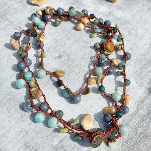 May include: A handmade necklace featuring a variety of beads in shades of blue, green, and tan, strung on a brown cord. The necklace is adorned with small, natural-looking stones and a decorative clasp. A starfish is in the background.