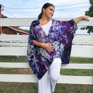 Op de afbeelding: Een vrouw draagt een paars-blauwe tie-dye kimono met een witte broek en sandalen. Ze staat voor een wit hek.